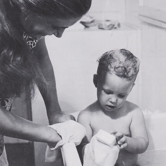 Kleinkind in der Badewanne hält ein Tuch und Seife in der Hand. Eine weibliche Erwachsene steht lächelnd und aufmerksam daneben.