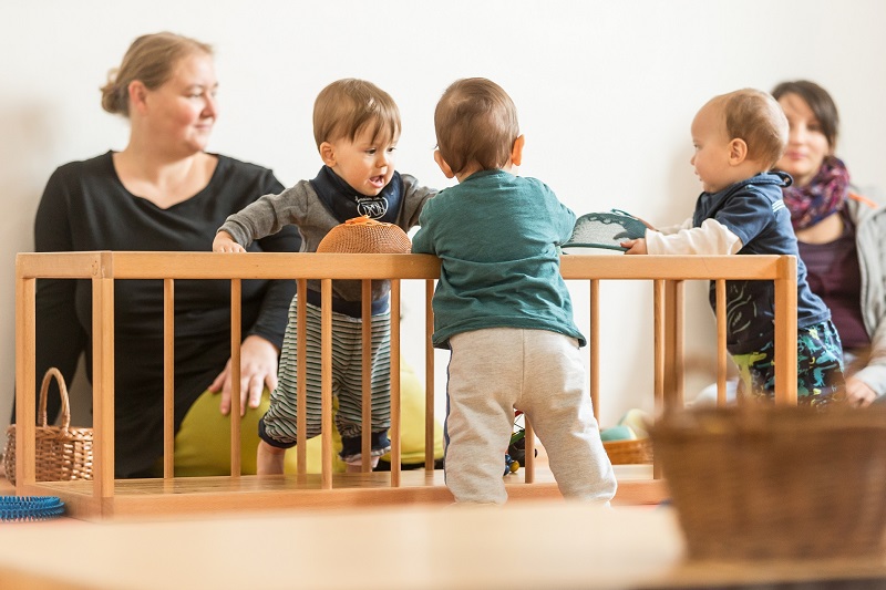 Im Pikler-SpielRaum: drei Babys stehen beim Pikler-Labyrint, im HIntergrund die Pädagogin Daniela Scheurer und eine beobachtende Mama