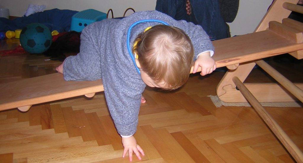 Kleinkind klettert über eine Rutsche und stützt sich mit den Zehen ab. Mit der rechten Hand tastet es nach dem Boden. 