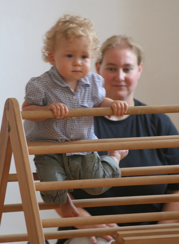 Kleinkind setzt sich auf eine Sprosse auf dem Pikler-Dreieck. Pädagogin sitzt dahinter