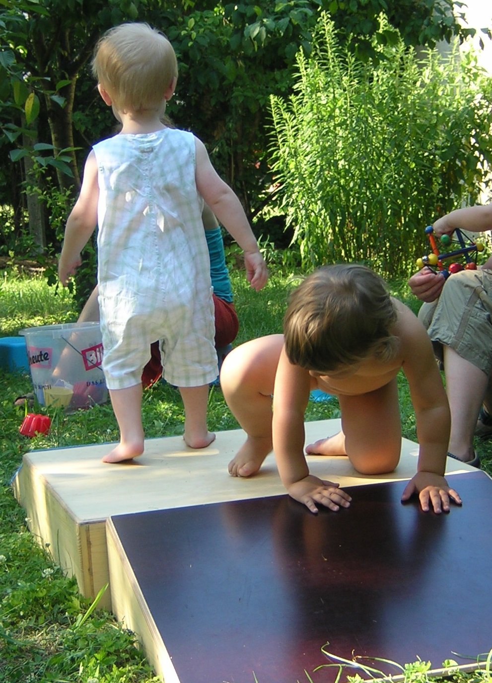 Ein Kleinkind steht auf einem kleinen Holzpodest mit schiefer rutschiger Ebene. Ein anderes Kleinkind will gerade die schiefe Ebene runterkrabbeln