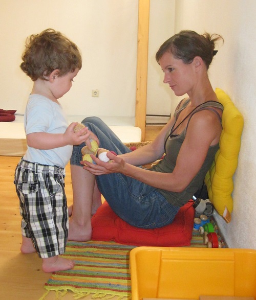 Im Pikler-SpielRaum: Ein Kleinkind bringt Bälle zu seiner Mutter, die am Boden sitzt und die Hände aufhält. Das Kleinkind legt die Bälle in die Hände der Mutter